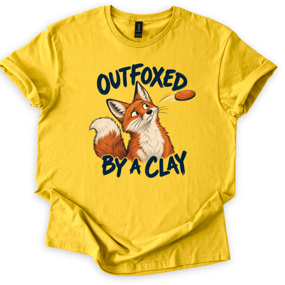Outfoxed by a Clay Comfort Colors T-shirt in Daisy, laid flat. A cartoon red fox looks up warily at an orange clay target flying past, between the dark navy brush letters OUTFOXED and BY A CLAY. The shirt has rolled sleeves and lies on a white background.