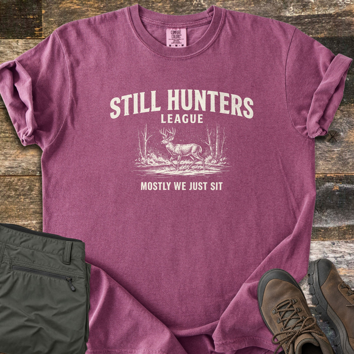 Still Hunters League Comfort Colors T-shirt in Berry, laid flat. Cream print of the words Still Hunters League above a sketched buck deer standing in grass between bare trees, with Mostly We Just Sit below. Sleeves rolled, on weathered wood with olive pants and brown hiking boots.