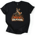 Rut Season Comfort Colors T-shirt in Black, laid flat. A cartoon buck in a helmet rides a green ATV through splashing mud above the words Rut Season in orange and cream mud-spattered letters. The shirt has rolled sleeves on a white background.