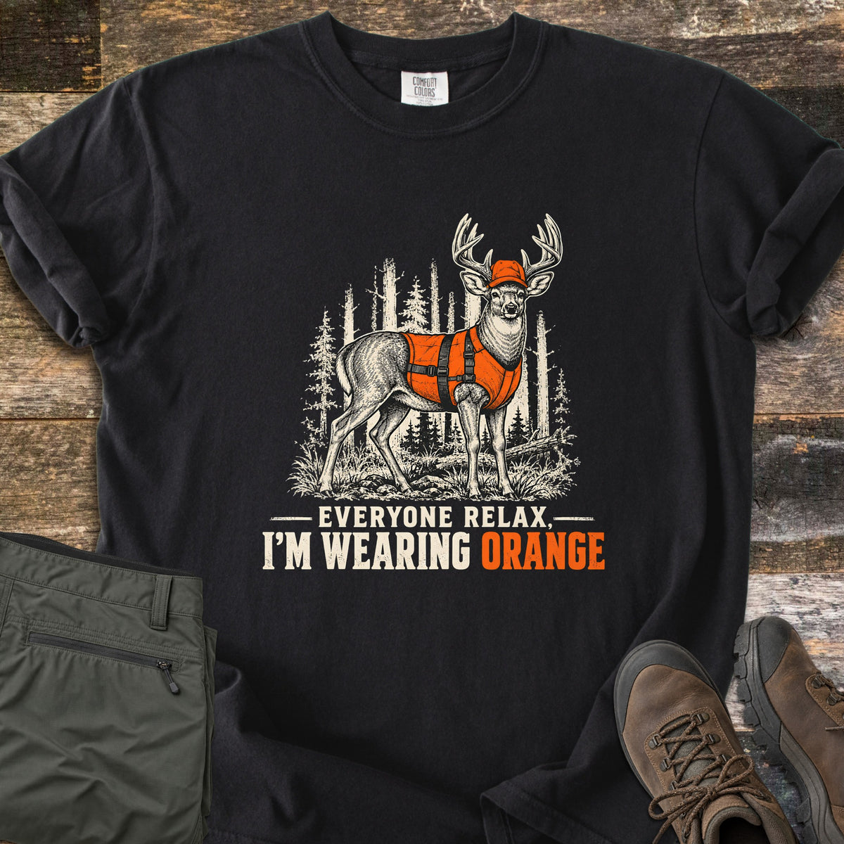 Everyone Relax, I'm Wearing Orange Comfort Colors T-shirt in Black, laid flat. A vintage-style deer with antlers wears an orange cap and orange vest in a pine forest, above the words Everyone Relax, I'm Wearing Orange. Olive pants and brown hiking boots lie on weathered wood.