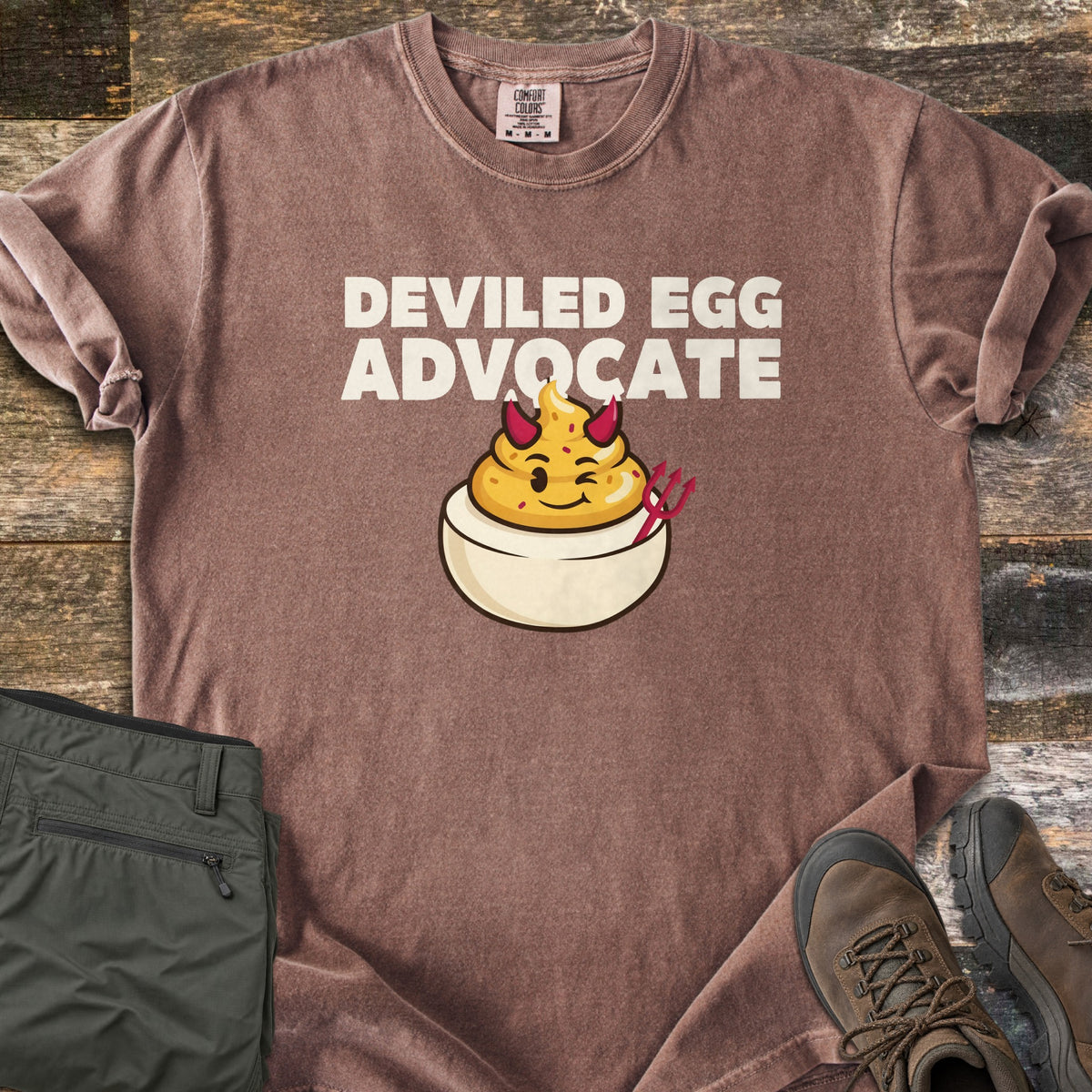 Deviled Egg Advocate Comfort Colors T-shirt in Espresso, laid flat. Cream letters read Deviled Egg Advocate above a winking cartoon deviled egg with red devil horns and a red pitchfork. Folded olive hiking pants and brown hiking boots sit on weathered wood planks.