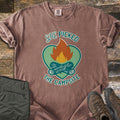 She Picked The Campsite Comfort Colors T-shirt in Espresso, laid flat. A campfire with crossed logs sits on a green heart, with the words She Picked above and The Campsite below. Sleeves are rolled on a rustic wood floor beside folded olive hiking pants and brown hiking boots.