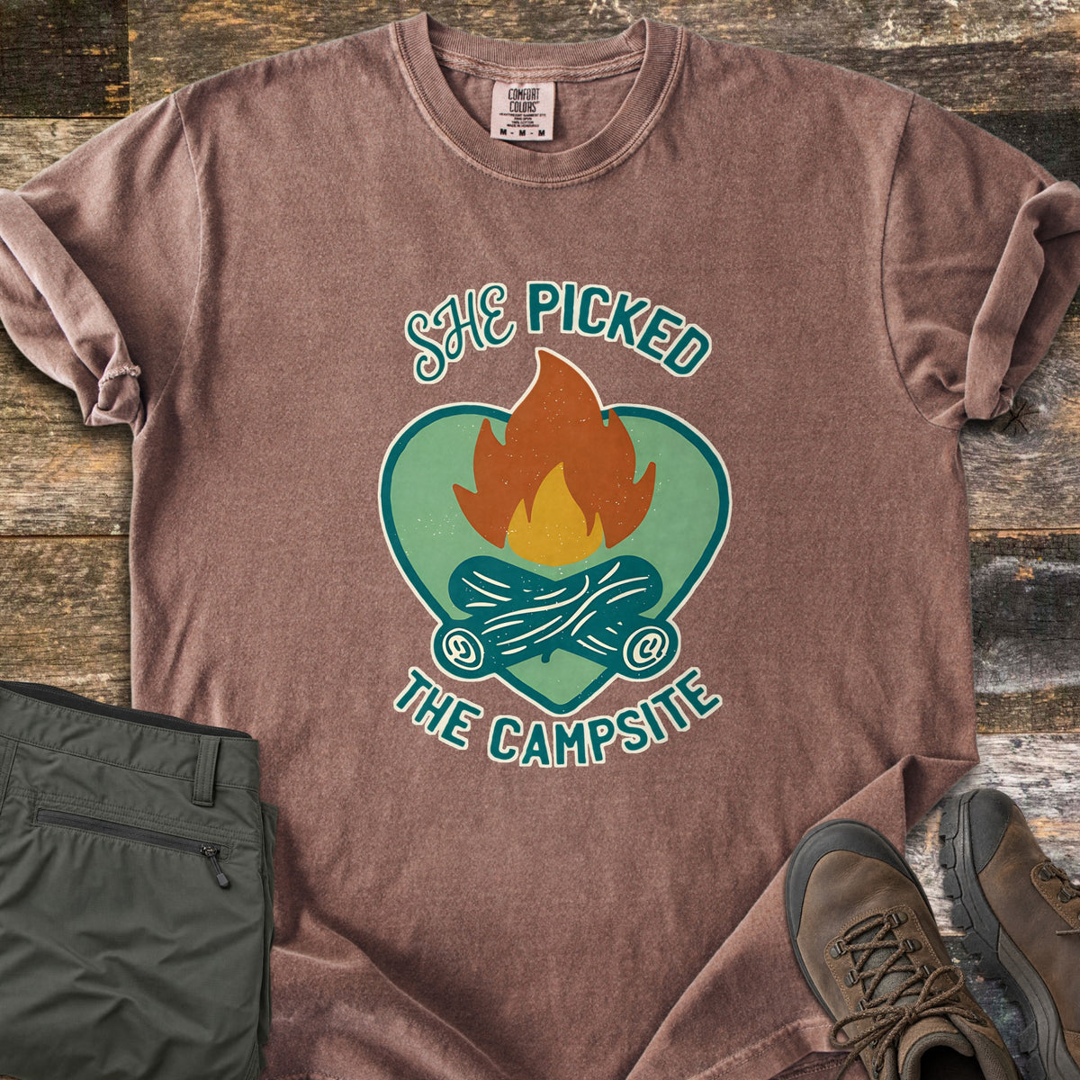 She Picked The Campsite Comfort Colors T-shirt in Espresso, laid flat. A campfire with crossed logs sits on a green heart, with the words She Picked above and The Campsite below. Sleeves are rolled on a rustic wood floor beside folded olive hiking pants and brown hiking boots.