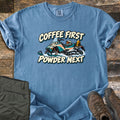Coffee First Powder Next Comfort Colors T-shirt in Blue Jean, laid flat. A cartoon snowmobile with a helmet on the seat and a yellow thermos strapped on back kicks up snow between the words Coffee First Powder Next. Olive hiking pants and brown boots sit on a wood floor.