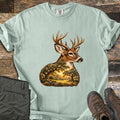 Whitetail Oak Grove Comfort Colors T-shirt in Bay, laid flat. A whitetail buck with branching antlers, its neck and chest filled with a sunlit grove of oak trees and grassy meadow. The shirt lies on weathered wood planks beside folded olive pants and brown hiking boots.