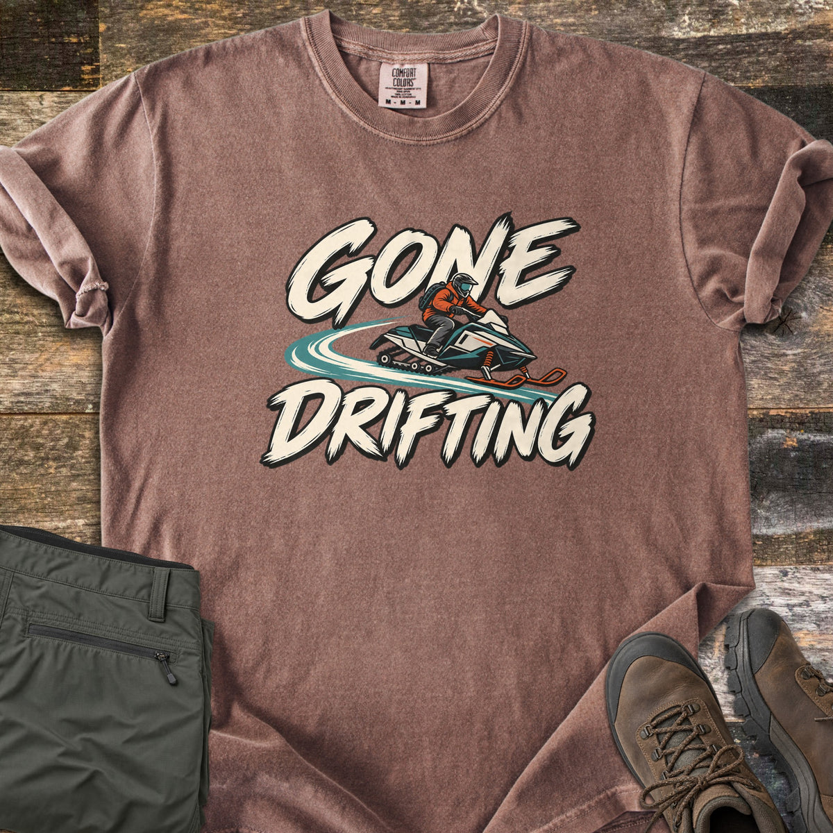 Gone Drifting Comfort Colors T-shirt in Espresso, laid flat. Cream brush letters read Gone Drifting around a rider in an orange jacket and helmet on a teal snowmobile with a swirling snow trail. On weathered wood beside folded olive pants and brown hiking boots.