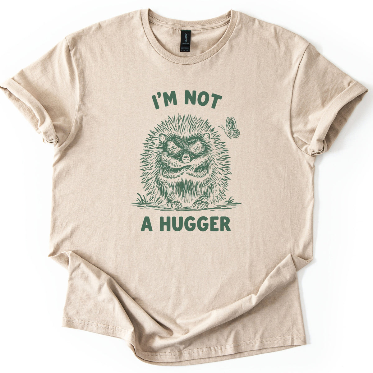 I'm Not A Hugger Comfort Colors T-shirt in Sand, laid flat. A scowling sketch-style hedgehog with crossed arms, a butterfly beside it, sits between the words I'm Not A Hugger in dark green. The sleeves are rolled on a white background.