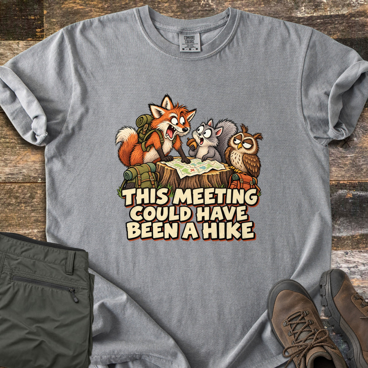 This Meeting Could Have Been a Hike Comfort Colors T-shirt in Grey, laid flat. A shouting cartoon fox, a squirrel holding an acorn and a sleepy owl gather at a trail map on a tree stump above the words This Meeting Could Have Been a Hike. Olive hiking pants and brown boots lie on wood.
