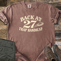 Back at 27 Trap Handicap Comfort Colors T-shirt in Espresso, laid flat. Cream print reads Back at 27 in an arch above Trap Handicap, with a clay target flying beside the 27. Sleeves rolled on a rustic wood surface, with olive cargo pants and brown hiking boots.