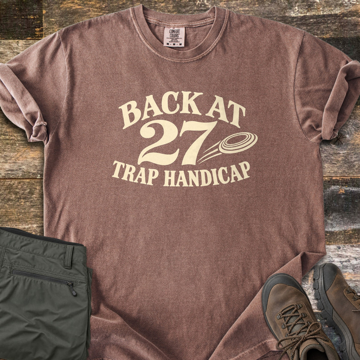 Back at 27 Trap Handicap Comfort Colors T-shirt in Espresso, laid flat. Cream print reads Back at 27 in an arch above Trap Handicap, with a clay target flying beside the 27. Sleeves rolled on a rustic wood surface, with olive cargo pants and brown hiking boots.