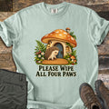 Please Wipe All Four Paws Comfort Colors T-shirt in Bay, laid flat. A hedgehog stands on a woven doormat outside a spotted orange mushroom house with a blue door, lantern and ferns, above the words Please Wipe All Four Paws. Gray pants and brown hiking boots lie on weathered wood.