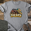 The GPS Has Questions Comfort Colors T-shirt in Grey, laid flat. A muddy orange side-by-side off-road vehicle climbs a rocky dirt trail past a wooden signpost and pine trees, above the words The GPS Has Questions. Sleeves are rolled on a wood floor beside olive pants and brown boots.