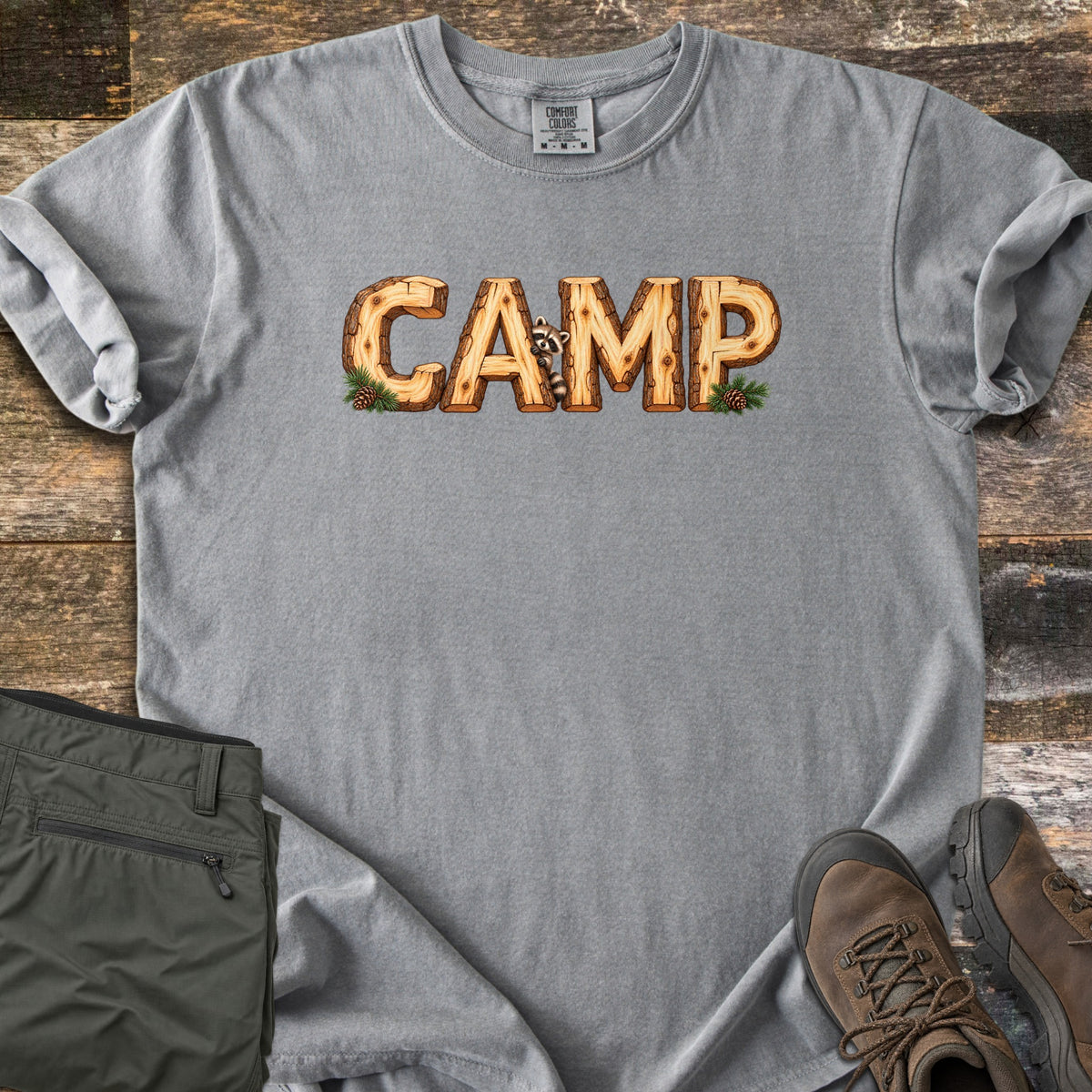 Camp Comfort Colors T-shirt in Grey, laid flat. The word CAMP is printed in letters made of cut wooden logs, with a raccoon peeking out between the A and M and pine sprigs with pinecones at each end. Olive pants and brown hiking boots sit on weathered wood planks.