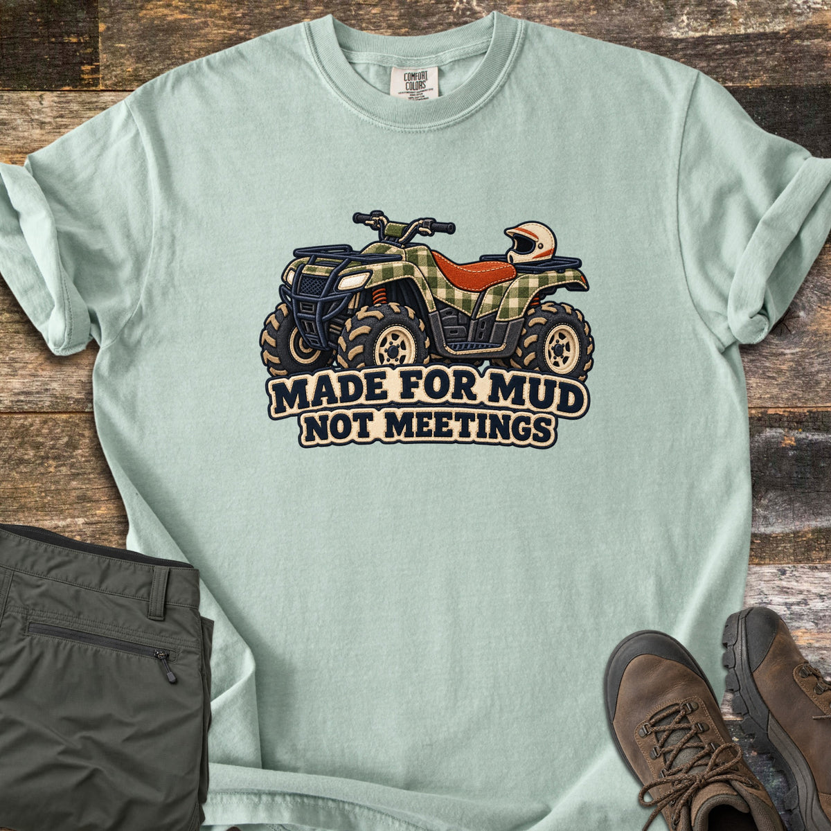Made for Mud Not Meetings Comfort Colors T-shirt in Bay, laid flat. A green plaid ATV with an orange seat, muddy tires and a white helmet on the rack sits above the words Made for Mud Not Meetings. It lies on weathered wood beside folded olive pants and brown hiking boots.