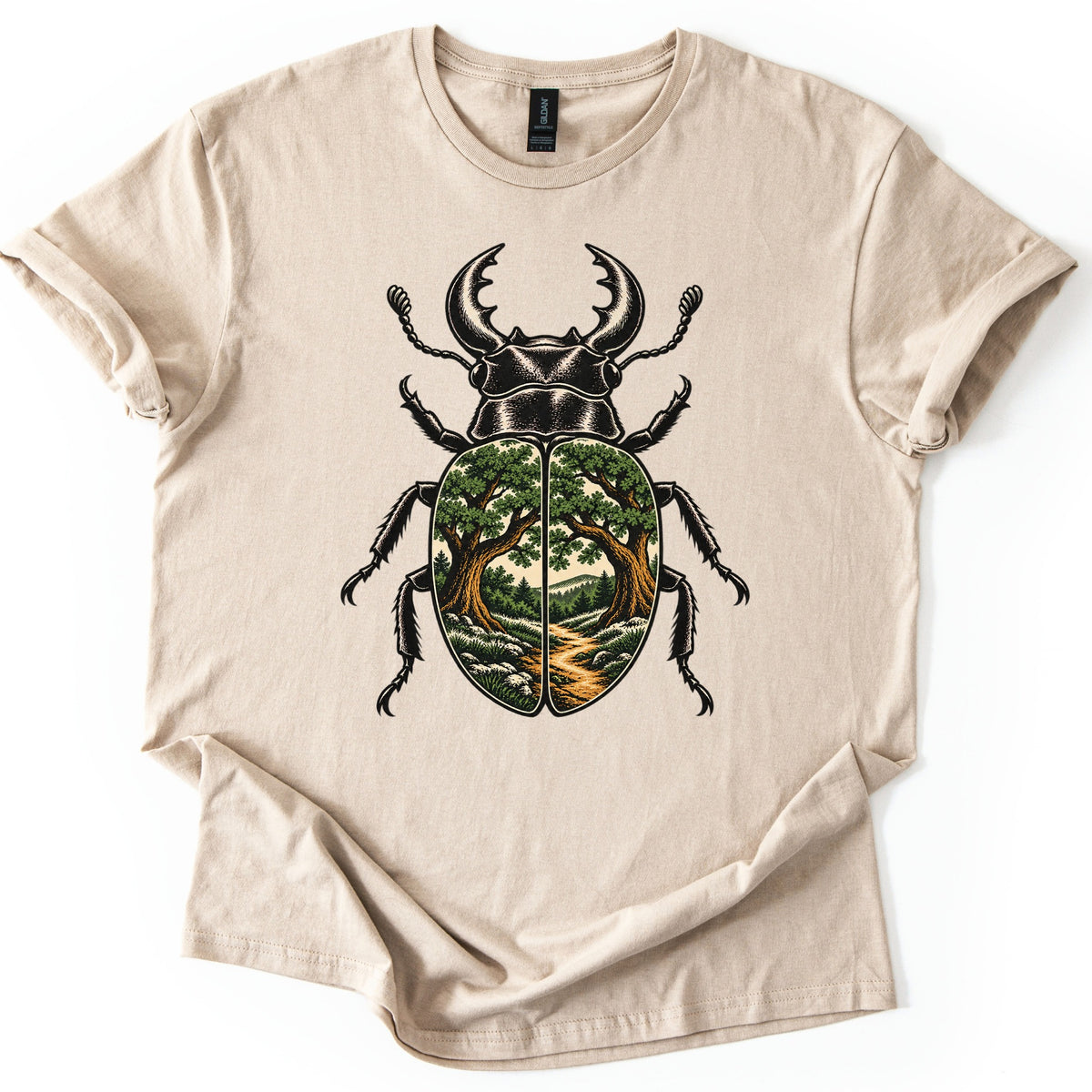 Beetle Forest Path Comfort Colors T-shirt in Sand, laid flat. A black ink stag beetle with curved pincers and spiny legs, its shell showing oak trees framing a dirt path through a green forest. No words are printed. The shirt lies on white with cuffed sleeves.