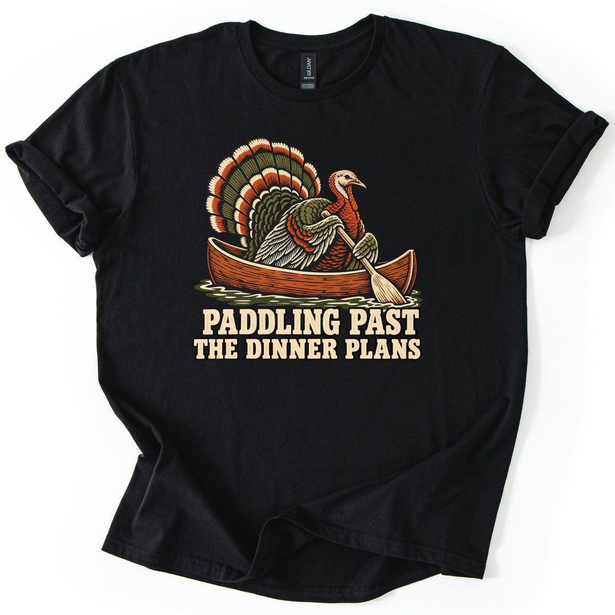 Paddling Past the Dinner Plans Comfort Colors T-shirt in Black, laid flat. A wild turkey with a fanned tail rows a wooden canoe across the water with a paddle, above the words Paddling Past the Dinner Plans in cream letters. The shirt lies on a white background with its sleeves rolled.