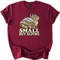 Small But Alpine Comfort Colors T-shirt in Maroon, laid flat. A pika sits on rocks, its body filled with a scene of snowy peaks, a lake and purple and pink wildflowers, above the words SMALL BUT ALPINE in cream letters. The shirt lies on white with rolled sleeves.