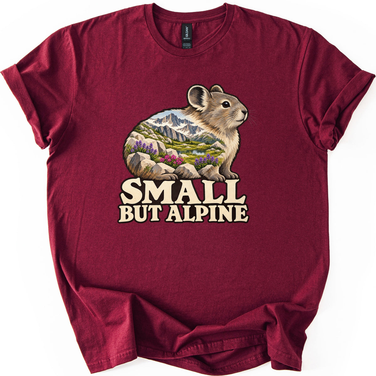 Small But Alpine Comfort Colors T-shirt in Maroon, laid flat. A pika sits on rocks, its body filled with a scene of snowy peaks, a lake and purple and pink wildflowers, above the words SMALL BUT ALPINE in cream letters. The shirt lies on white with rolled sleeves.