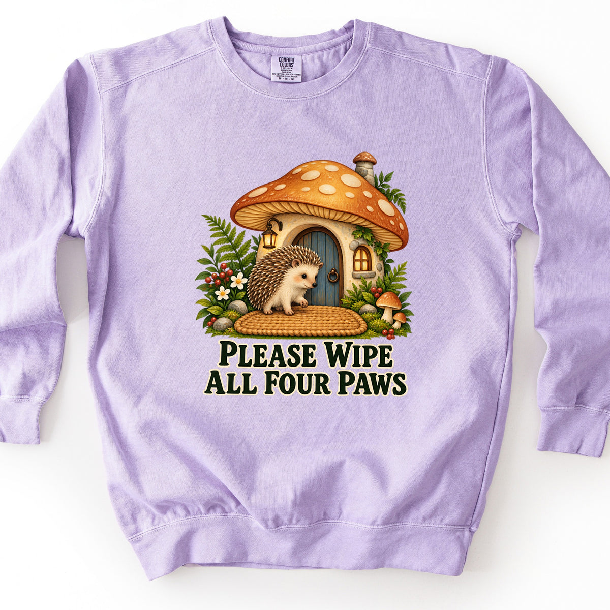Please Wipe All Four Paws Comfort Colors crewneck sweatshirt in Orchid, laid flat. A hedgehog stands on a woven doormat outside an orange-capped mushroom house with a blue door, lantern, ferns, and berries, above the words Please Wipe All Four Paws in dark green. Shown on a white background.