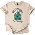 Better When I'm Climbing Comfort Colors T-shirt in Sand, laid flat. The words Life is better when arch over a window scene of a person facing a teal mountain with a winding trail, a yellow sun and two pine trees, with I'm Climbing below. Sleeves cuffed, on a white surface.