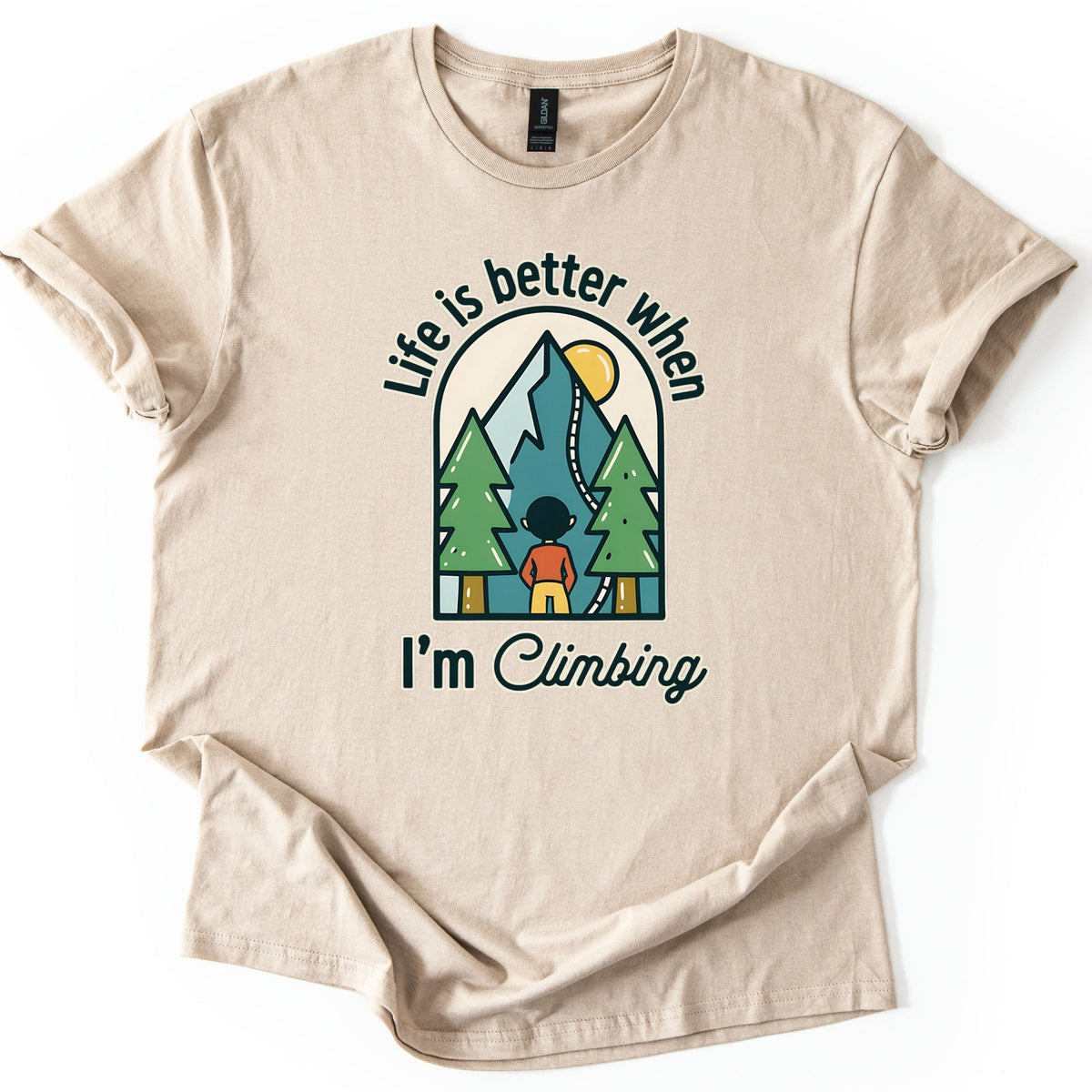 Better When I'm Climbing Comfort Colors T-shirt in Sand, laid flat. The words Life is better when arch over a window scene of a person facing a teal mountain with a winding trail, a yellow sun and two pine trees, with I'm Climbing below. Sleeves cuffed, on a white surface.