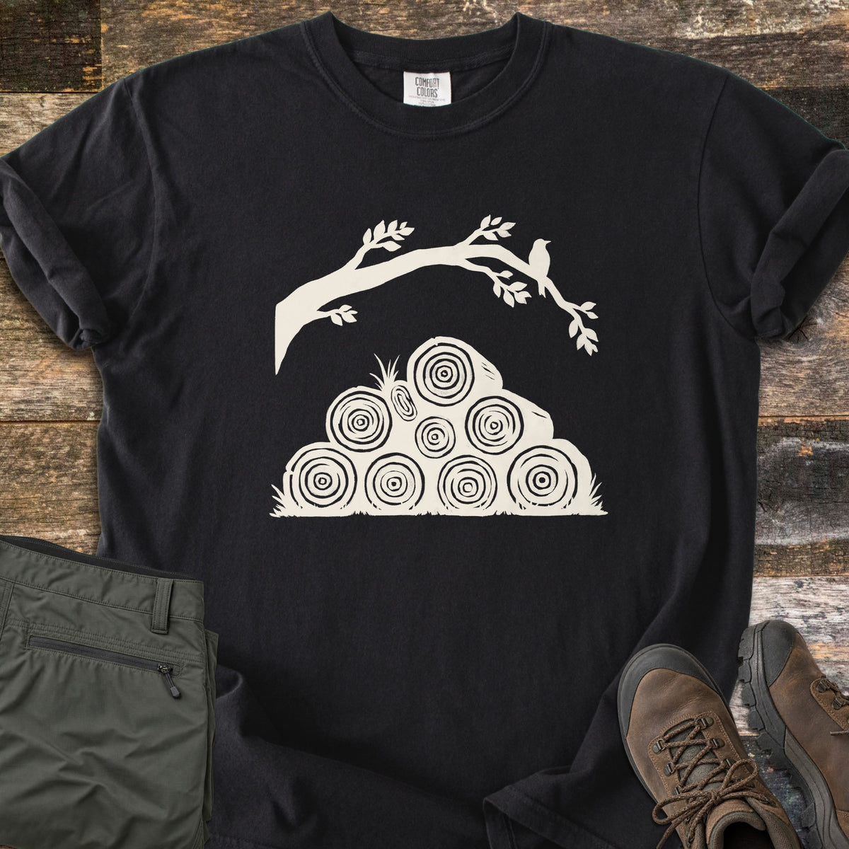 Stacked Firewood Comfort Colors T-shirt in Black, laid flat. An ivory print of a leafy tree branch with a small bird perched on it, arching over a pyramid of stacked firewood logs showing tree rings, with no words. Olive cargo pants and brown hiking boots sit on weathered wood planks.