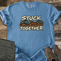 Stuck Together Comfort Colors T-shirt in Blue Jean, laid flat. Two teal ATVs stuck in brown mud, one towing the other with an orange strap, between the words Stuck Together in cream brush lettering. Olive pants and brown hiking boots lie beside it on weathered wood.