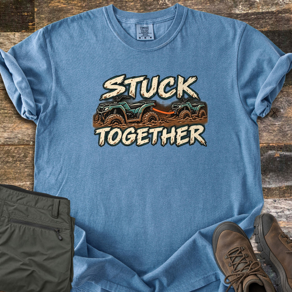 Stuck Together Comfort Colors T-shirt in Blue Jean, laid flat. Two teal ATVs stuck in brown mud, one towing the other with an orange strap, between the words Stuck Together in cream brush lettering. Olive pants and brown hiking boots lie beside it on weathered wood.