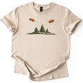 Crossing Clays Comfort Colors T-shirt in Sand, laid flat. Two orange clay targets fly on crossing paths with motion trails above three dark green pine trees on a grassy hill, with no words. The shirt lies on a white background with cuffed sleeves.