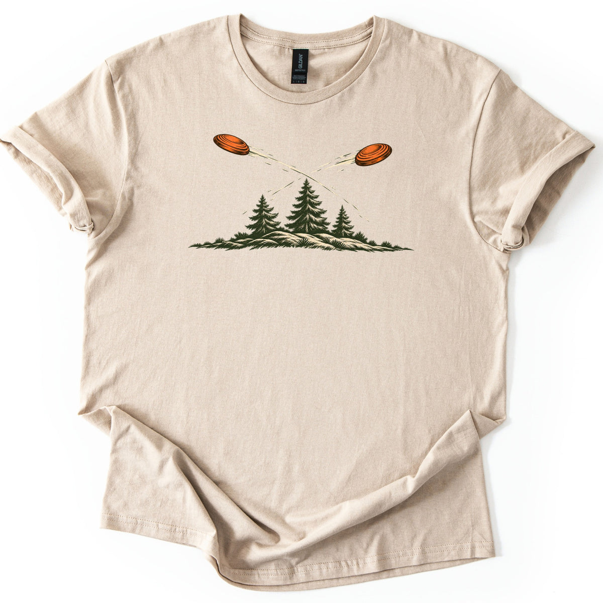 Crossing Clays Comfort Colors T-shirt in Sand, laid flat. Two orange clay targets fly on crossing paths with motion trails above three dark green pine trees on a grassy hill, with no words. The shirt lies on a white background with cuffed sleeves.