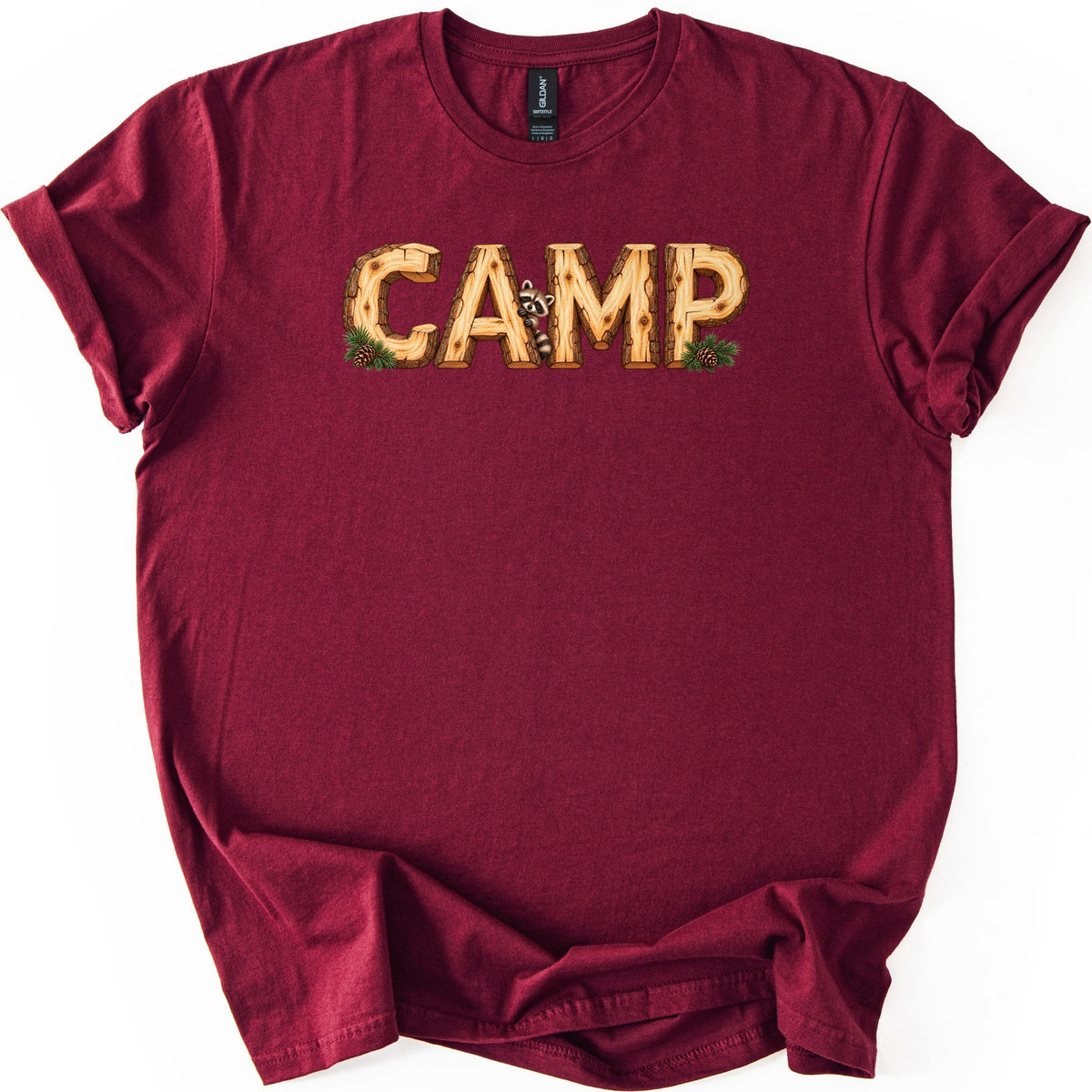 Camp Comfort Colors T-shirt in Maroon, laid flat. The word CAMP is printed in letters made of cut logs with bark edges, a small raccoon peeking out between the A and M, and pine sprigs with pinecones at the corners. The sleeves are cuffed on a white background.