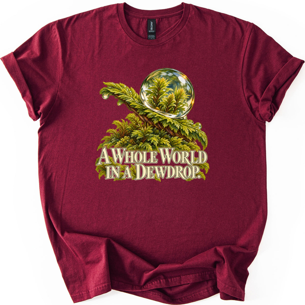 A Whole World in a Dewdrop Comfort Colors T-shirt in Maroon, laid flat. A clear dewdrop rests on a feathery green moss frond, showing tiny plants inside it, above the cream words A Whole World in a Dewdrop. The shirt lies on a white surface with sleeves rolled.