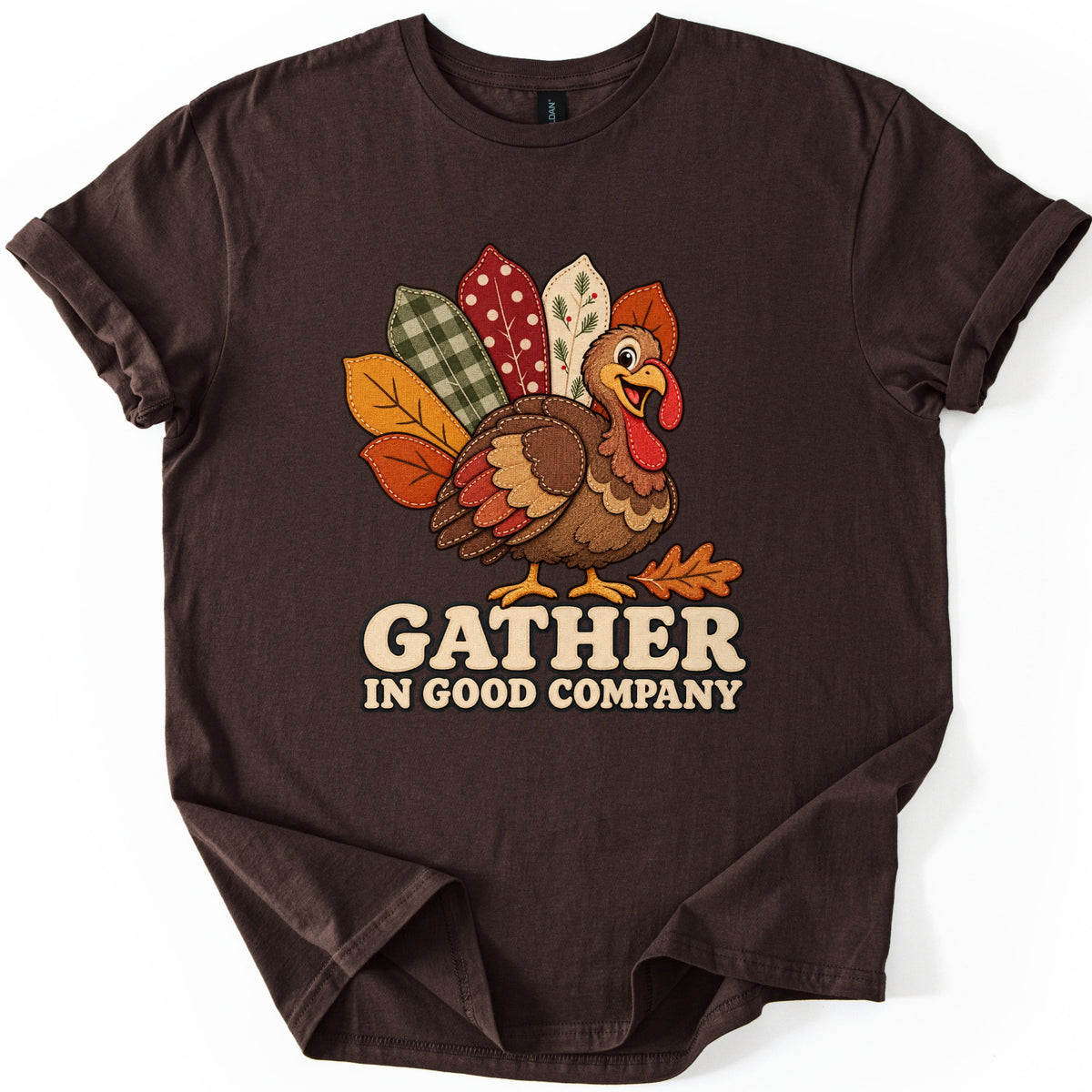 Gather in Good Company Comfort Colors T-shirt in Dark Chocolate, laid flat. A smiling cartoon turkey with stitched patchwork tail feathers in plaid, polka dot and autumn leaf prints stands above the words GATHER IN GOOD COMPANY in cream letters. Sleeves cuffed, on a white background.