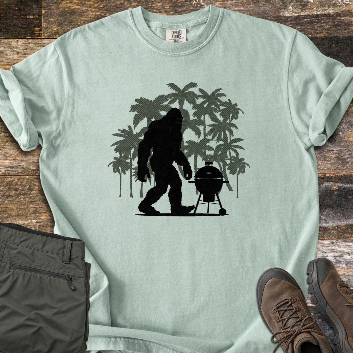 Squatch BBQ Smoker Comfort Colors T-shirt in Bay, laid flat. A black Bigfoot silhouette walks past a kettle grill on wheels, set against dark green palm tree silhouettes; no words are printed. The shirt lies on weathered wood beside olive pants and brown hiking boots.