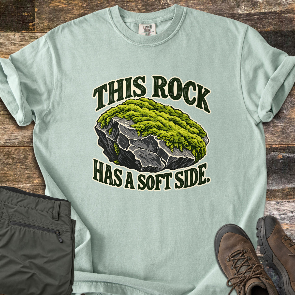 This Rock Has a Soft Side Comfort Colors T-shirt in Bay, laid flat. An illustrated gray boulder topped with thick green moss sits between the words This Rock above and Has a Soft Side. below. Olive hiking pants and brown hiking boots lie beside it on weathered wood planks.