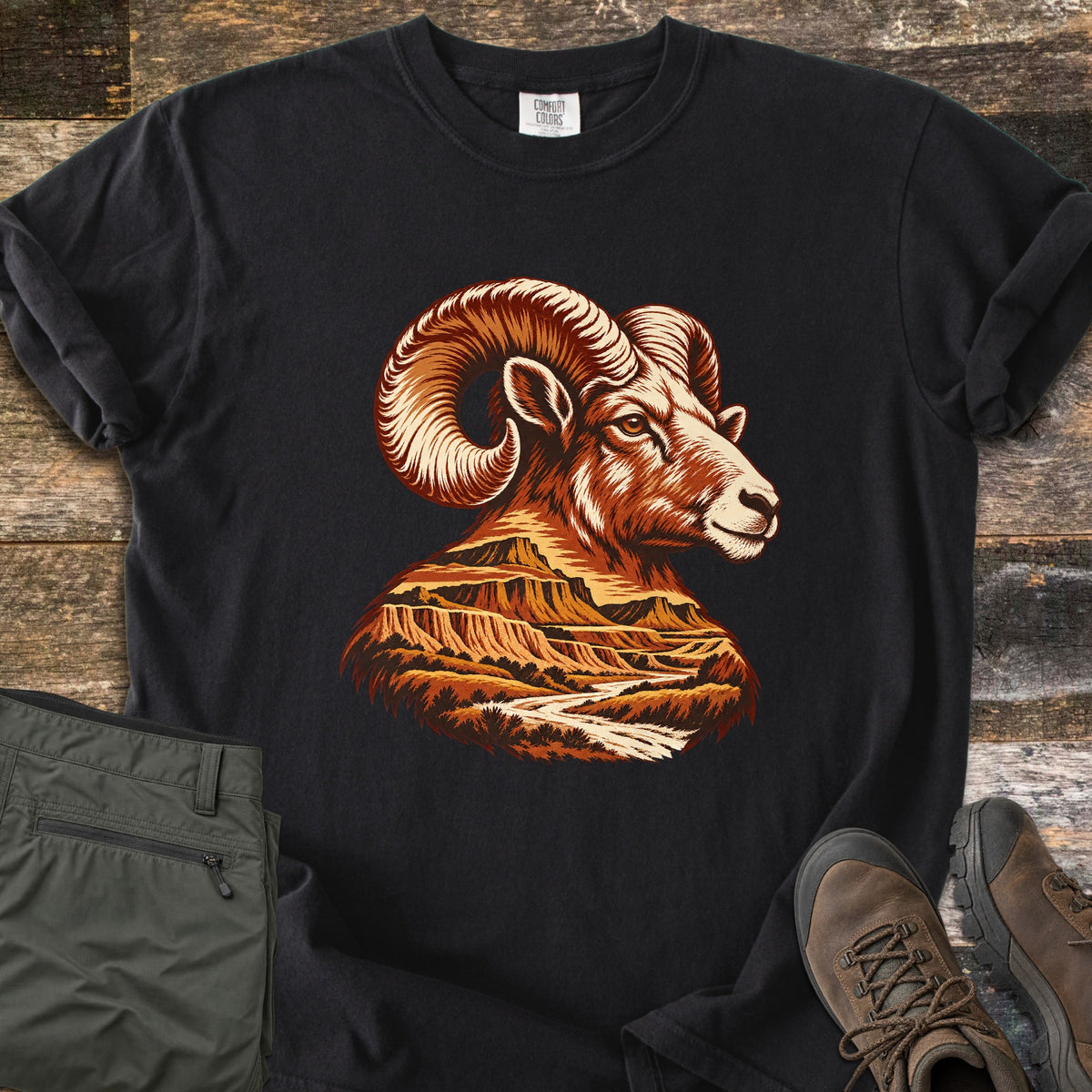Bighorn Badlands Comfort Colors T-shirt in Black, laid flat. A bighorn ram's head in rust and cream tones, its neck and chest filled with a badlands scene of buttes and a winding river. Shown on weathered wood with olive pants and brown hiking boots.
