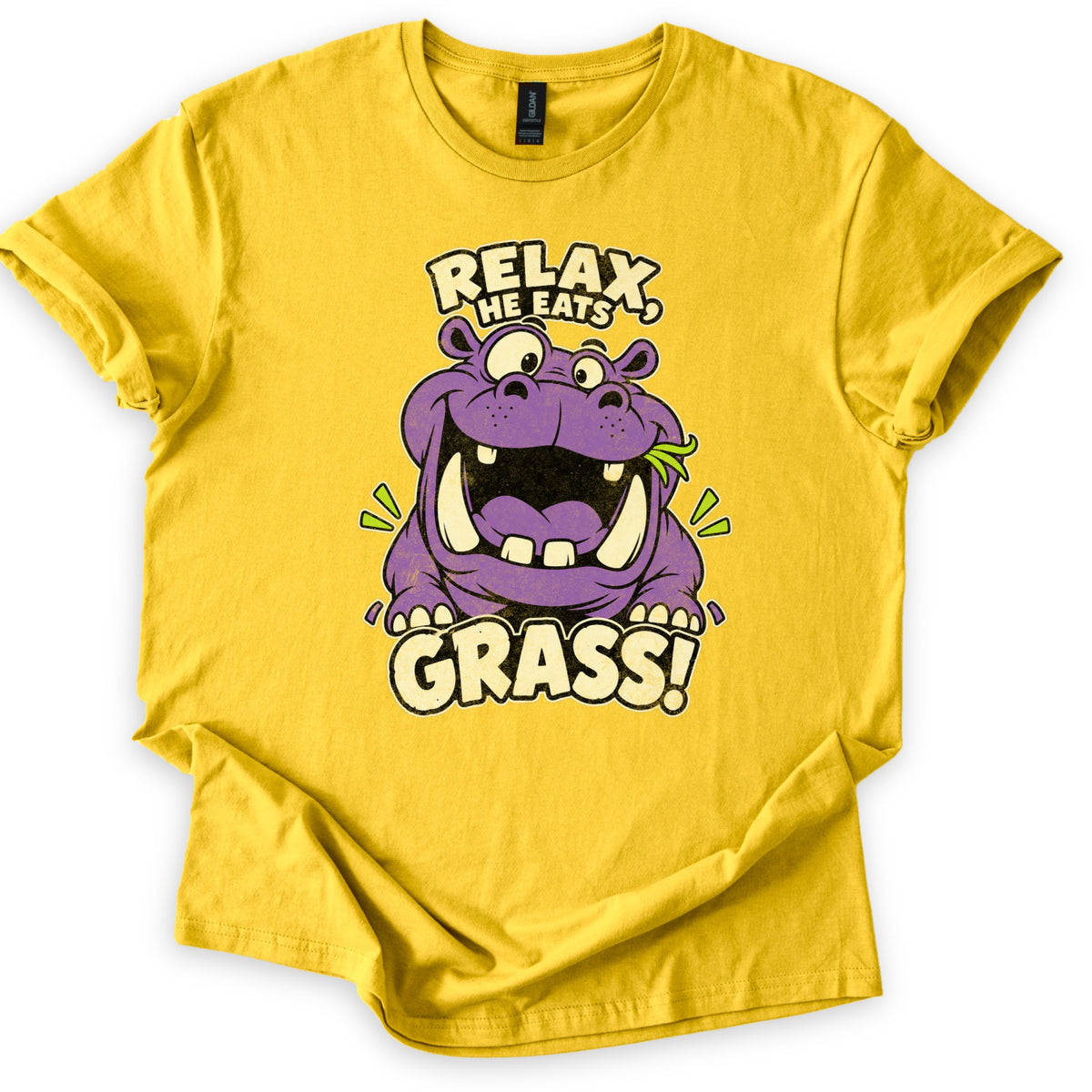 Relax He Eats Grass Comfort Colors T-shirt in Daisy, laid flat. A grinning purple cartoon hippo with big tusks and a blade of grass in its mouth sits between the words RELAX, HE EATS and GRASS! in cream letters. The shirt lies on white with rolled sleeves.