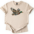 Mallard Flooded Oaks Comfort Colors T-shirt in Sand, laid flat. A mallard drake flies across the chest, its wings and body filled with a scene of moss-draped trees standing in still floodwater under an orange and blue sky. No words. Sleeves rolled, on a white background.