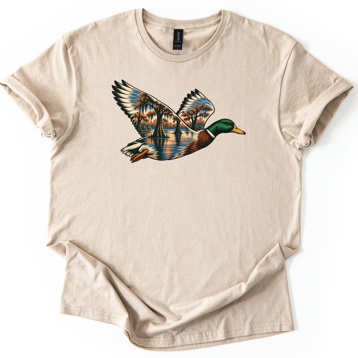 Mallard Flooded Oaks Comfort Colors T-shirt in Sand, laid flat. A mallard drake flies across the chest, its wings and body filled with a scene of moss-draped trees standing in still floodwater under an orange and blue sky. No words. Sleeves rolled, on a white background.