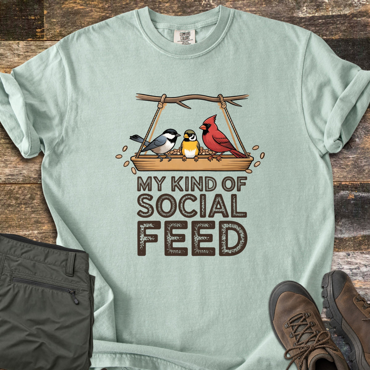 My Kind Of Social Feed Comfort Colors T-shirt in Bay, laid flat. A chickadee, a small yellow bird and a red cardinal sit on a rope-hung tray feeder full of seeds above the words My Kind Of Social Feed in brown. Olive hiking pants and brown hiking boots lie on weathered wood.