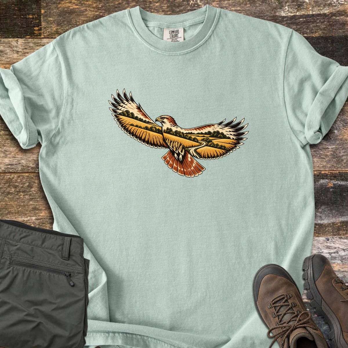 Hawk Over Golden Fields Comfort Colors T-shirt in Bay, laid flat. A hawk with outstretched wings and a rust-red tail, its wings filled with golden fields, green trees and a winding path; no words. Folded olive hiking pants and brown hiking boots sit beside it on weathered wood.