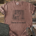 Someone Ate The Wreath Comfort Colors T-shirt in Espresso, laid flat. A sketched deer nibbles a wreath on a cabin's front door, framed by corner marks above the words Someone Ate The Wreath. Shown on rustic wood planks with folded olive pants and brown hiking boots.