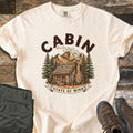 Cabin State Of Mind Comfort Colors T-shirt in Ivory, laid flat. A log cabin with a smoking stone chimney sits among pine trees and mountains under the arched word CABIN, with a banner reading State Of Mind. Green pants and brown hiking boots lie beside it on weathered wood.