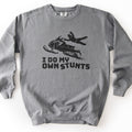 I Do My Own Stunts Comfort Colors crewneck sweatshirt in Grey, laid flat. A black print shows a rider in a helmet flung off an airborne snowmobile as snow sprays behind it, above the words I DO MY OWN STUNTS. The sweatshirt lies on a white surface.