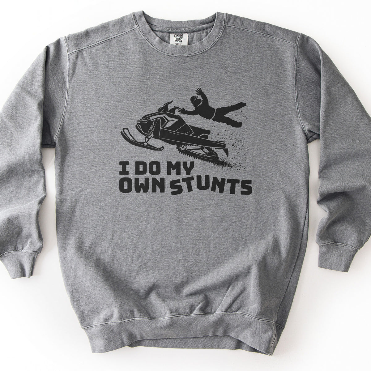 I Do My Own Stunts Comfort Colors crewneck sweatshirt in Grey, laid flat. A black print shows a rider in a helmet flung off an airborne snowmobile as snow sprays behind it, above the words I DO MY OWN STUNTS. The sweatshirt lies on a white surface.