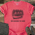 Who Borrowed The Canoe Comfort Colors T-shirt in Watermelon, laid flat. A black sketch of three otters in a canoe before pine trees, one holding a lantern, framed by camera corner marks above the words Who Borrowed The Canoe. Olive hiking pants and brown boots sit on dark wood.