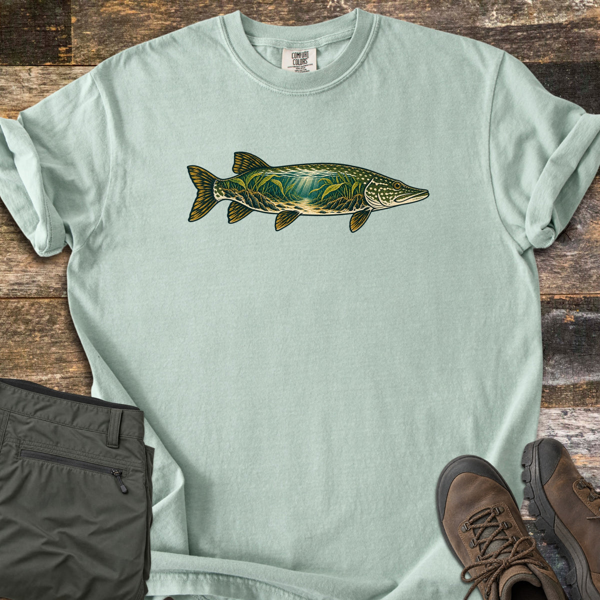 Pike Weed Bed Comfort Colors T-shirt in Bay, laid flat. A northern pike illustration across the chest, its body filled with an underwater scene of green weeds and light rays. The shirt lies on weathered wood beside folded olive pants and brown hiking boots.