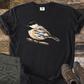 Chickadee Winter Creek Comfort Colors T-shirt in Black, laid flat. A black-capped chickadee perches on a snowy branch, its body filled with a winter scene of a creek, snowbanks and bare trees. The shirt lies on weathered wood beside olive pants and brown hiking boots.
