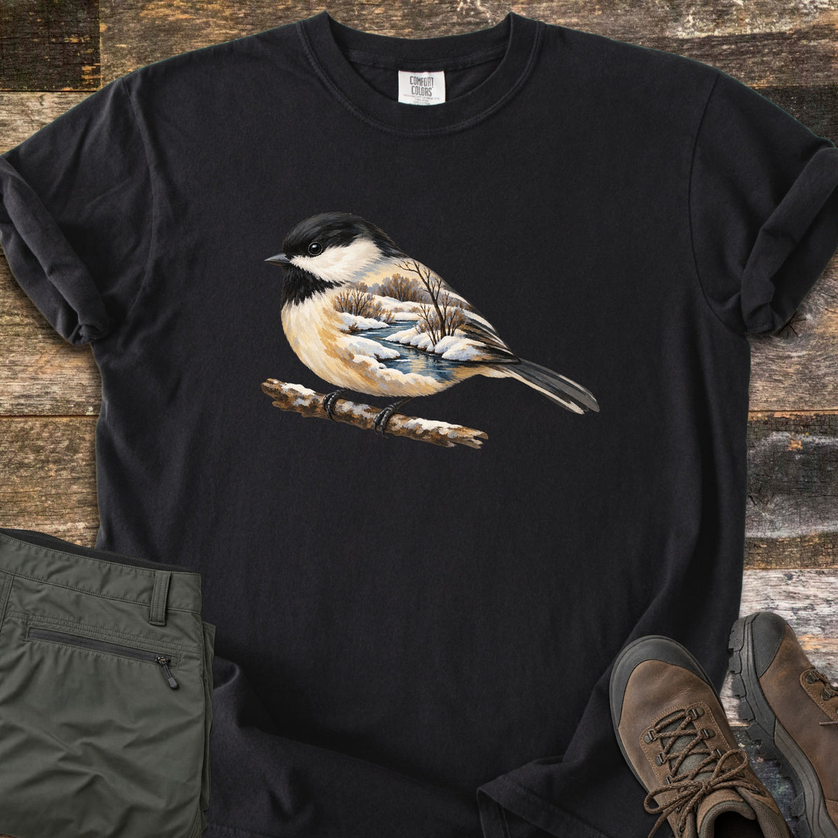 Chickadee Winter Creek Comfort Colors T-shirt in Black, laid flat. A black-capped chickadee perches on a snowy branch, its body filled with a winter scene of a creek, snowbanks and bare trees. The shirt lies on weathered wood beside olive pants and brown hiking boots.