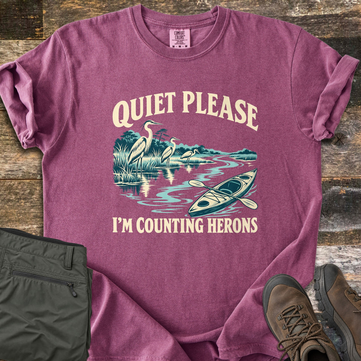 Counting Herons Comfort Colors T-shirt in Berry, laid flat. Printed text Quiet Please arches over three herons standing in marsh grass by a winding river with a kayak and paddle, above I'm Counting Herons. Olive cargo pants and brown hiking boots sit on weathered wood.